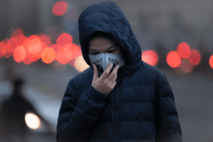 A person in a blue hooded jacket wears a face mask while standing in an urban setting with blurred red and white lights in the background. Their hand is touching the mask, and the atmosphere appears cold and overcast.