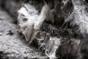 Close-up of fibrous, white asbestos fibers clinging to a rough, dark mineral surface. The fibers are dense and appear soft and wispy, contrasting with the hard texture of the mineral.