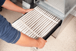 A person wearing a blue long-sleeve shirt is installing or removing a white pleated air filter from a HVAC system. The filter is being held with both hands above a speckled, light-colored floor.