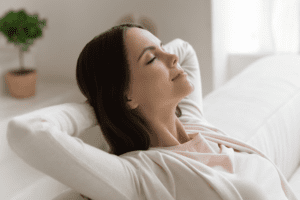 A woman with long brown hair is relaxing on a couch with her hands behind her head, eyes closed, and a peaceful expression. She is wearing a light sweater. A green potted plant is blurred in the background.