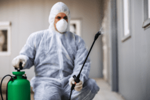 A person in protective gear and a face mask uses a spray wand connected to a green tank. They are spraying near a building, suggesting disinfection or pest control work. The background features gray walls and windows.