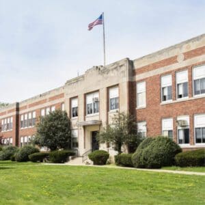 A large, brick school building with multiple windows sits surrounded by green lawns and shrubs. An American flag flies atop the building's central section under a partly cloudy sky, while concerns about asbestos in schools highlight the importance of safety and renovation.