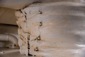 Close-up of a textured, weathered white pipe joint wrapped with fraying rope and asbestos seam tape, giving an aged and industrial appearance.