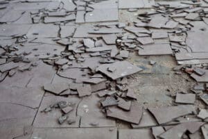 Broken and cracked asbestos floor tiles lie scattered across a dusty surface, creating a jagged pattern. The pieces vary in size and shape, revealing the underlying surface.