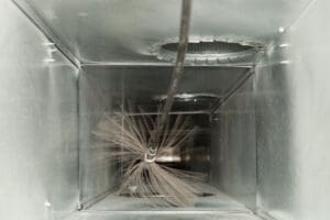 A duct cleaning brush rotates inside a metal air duct, emphasizing the importance of cleaning to ensure clean air ducts. The brush bristles blur as they spin, and the rectangular duct walls shine silver under the light.