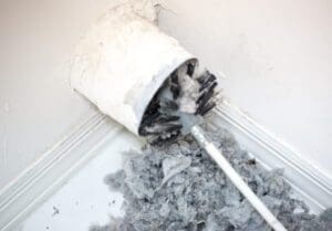 A white plastic vent in the wall showcases a brush inserted for duct cleaning, while a pile of gray lint and debris has gathered on the floor beneath it. The wall and baseboard are pristine white.