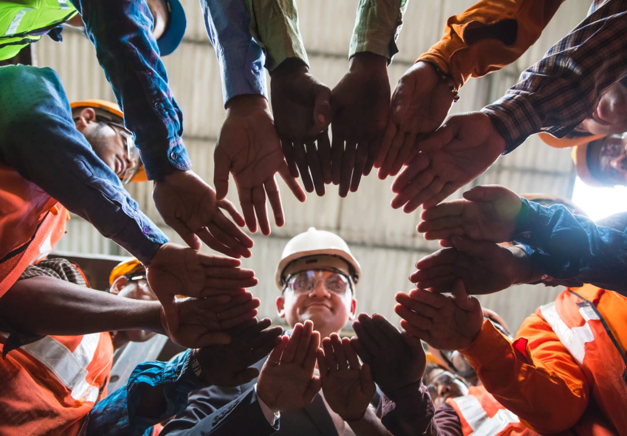 A group of workers wearing helmets and safety gear stand in a circle with their hands joined in the center, viewed from below. The background shows a corrugated metal roof. Their diverse clothing, perhaps suited for industrial cleaning, indicates different roles or trades.