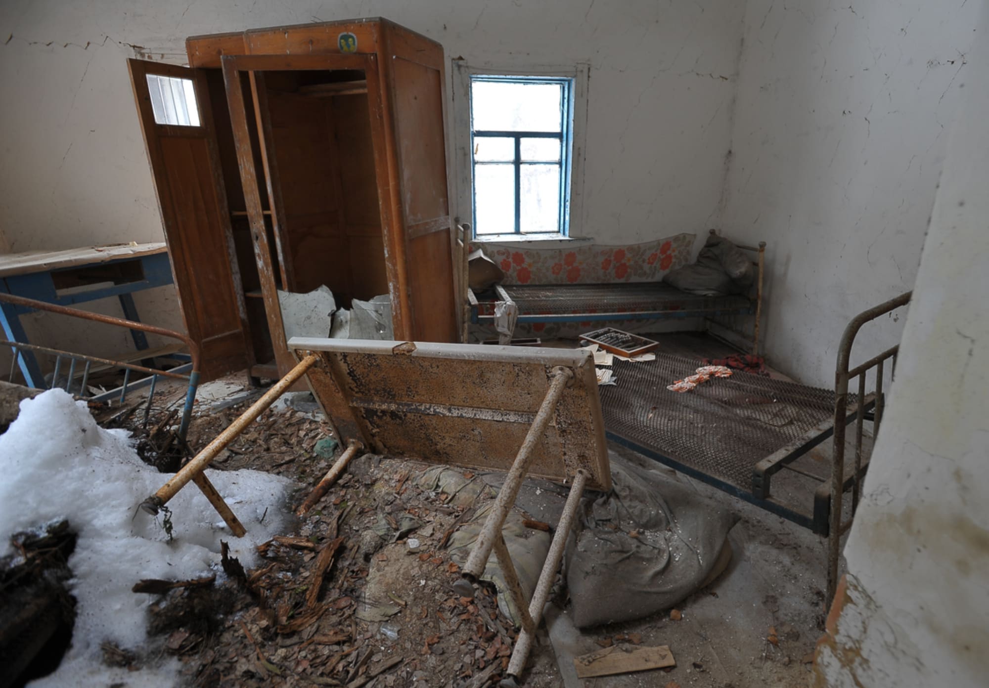 The room, in desperate need of interior renovation, features broken furniture—a bed, chair, and table—that sits amid debris and dirt. Light filters through a small window as snow melts by the entrance. The walls are cracked and peeling, emphasizing the urgent call for a transformation.