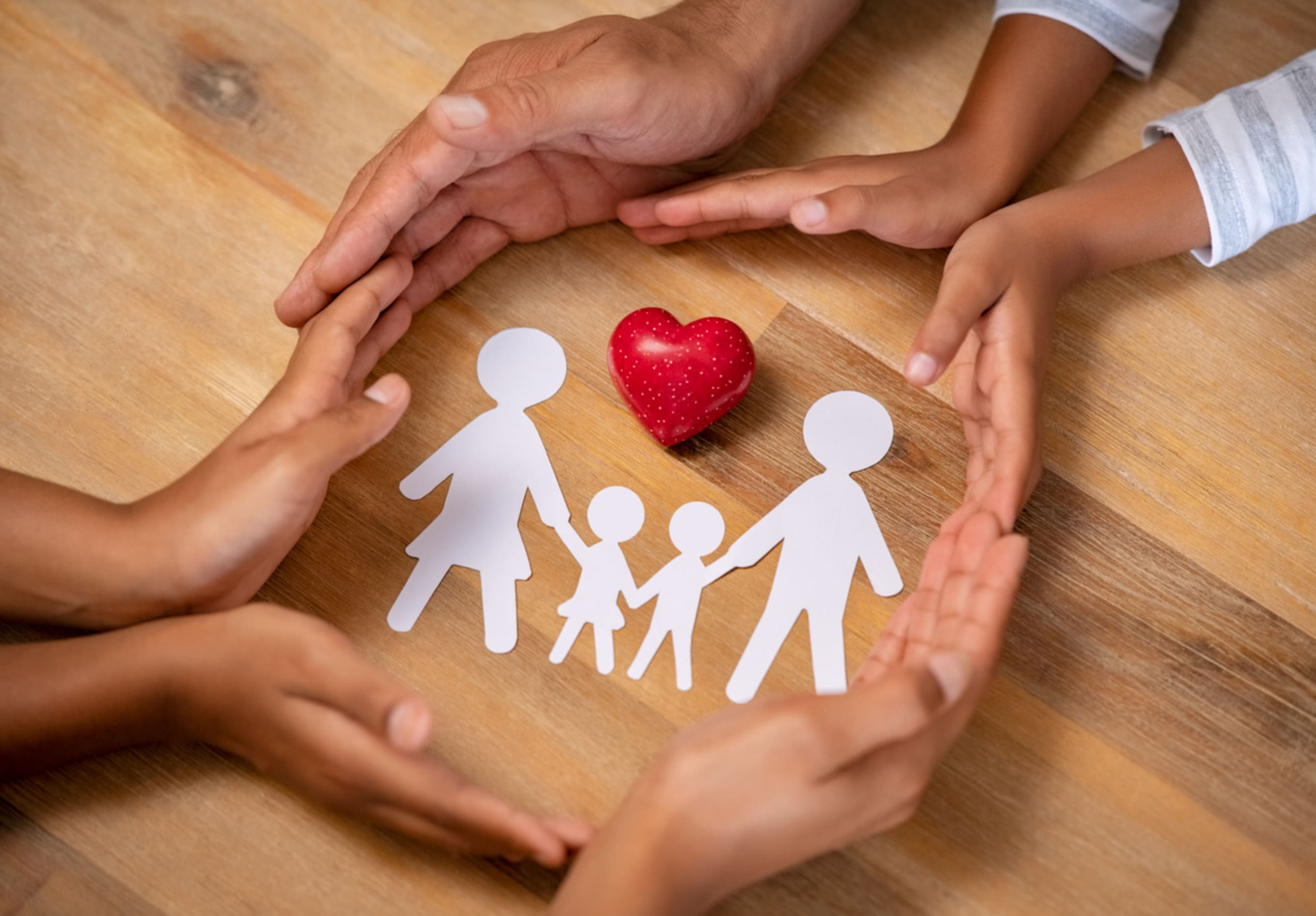 Hands of diverse individuals form a circle around paper cutouts of a family and a red heart on a wooden table. These cutouts represent two adults and two children, symbolizing love, unity, and the importance of safe environments akin to lead abatement in communities.