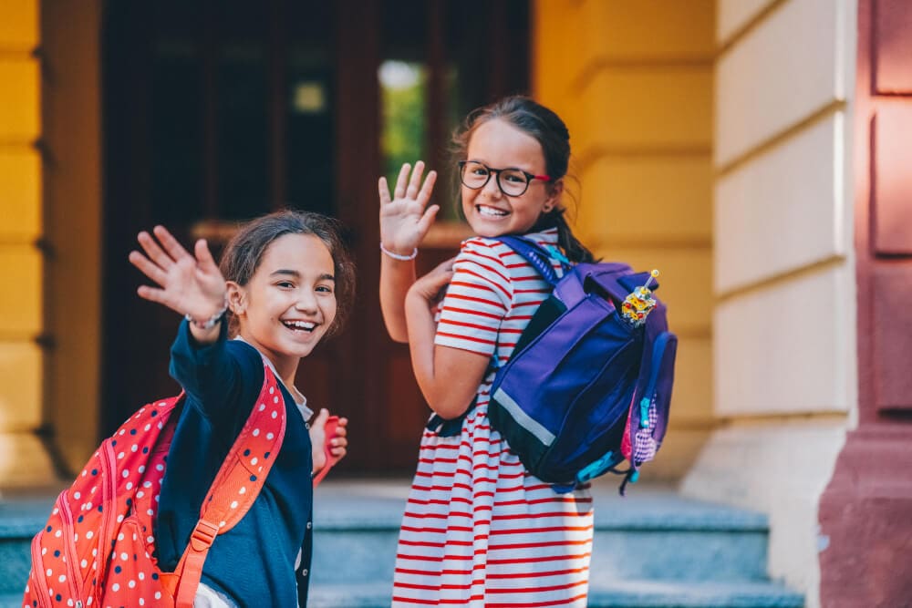 Two smiling girls with backpacks wave at the camera while standing on stone steps. The girl on the left wears a polka dot backpack, and the girl on the right, wearing glasses, has a purple backpack. They're outside a building with yellow and red walls, where duct cleaning is taking place.