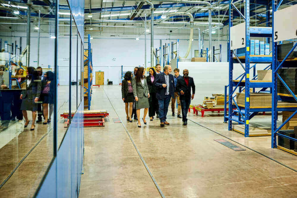 A group of people dressed in business attire walk through a large industrial warehouse, discussing industrial maintenance strategies. The space has high ceilings, shelves with various items, and a reflective glass wall on the left, all kept spotless by top-tier cleaning services.