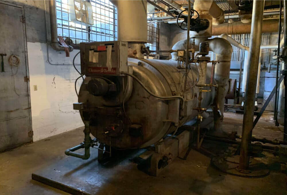 A large industrial boiler is situated in a dimly lit mechanical room, awaiting interior demolition. The metal structure features various pipes and valves, with signs of wear and rust. Walls and ceiling are constructed from concrete and metal beams, adding to the utilitarian atmosphere.