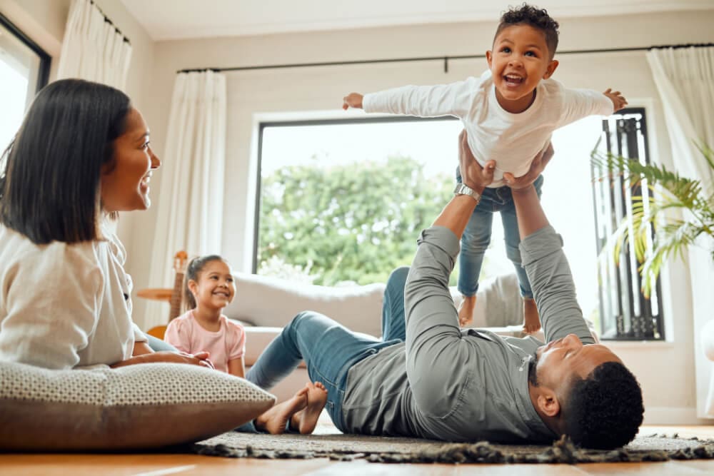 A family enjoys a playful moment in a bright living room. A man lies on the floor, lifting a laughing child into the air. Nearby, a woman and young girl sit smiling, fresh from cleaning chores. Large windows with curtains open to a view of greenery.