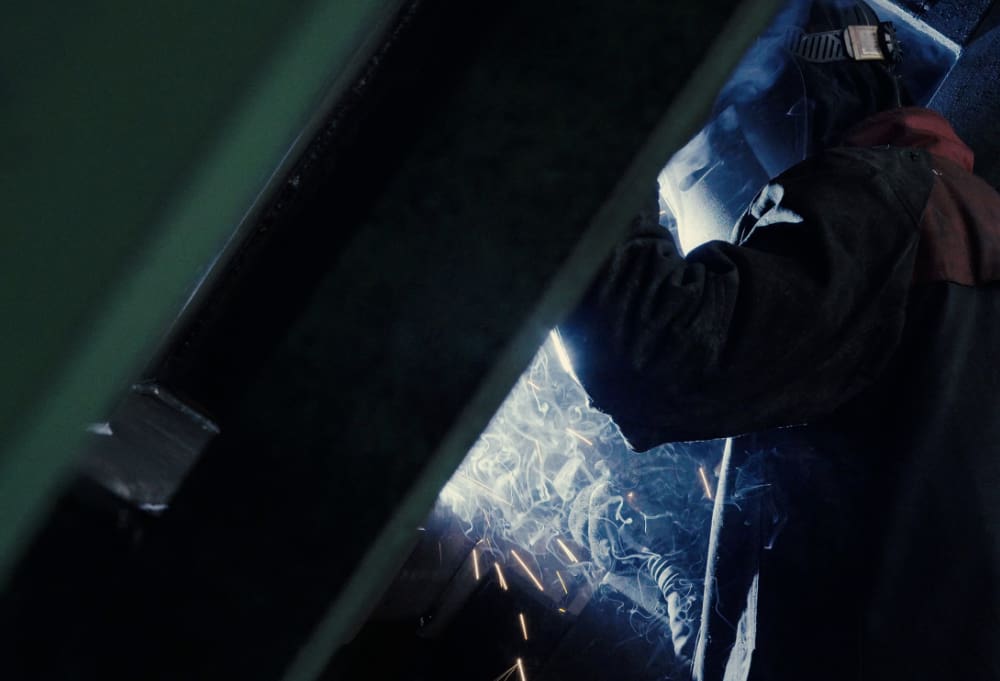 A welder wearing protective gear skillfully works on metal amidst the chaos of interior demolition. Sparks fly and smoke rises around the welding area, partially obscured by a large metal frame.