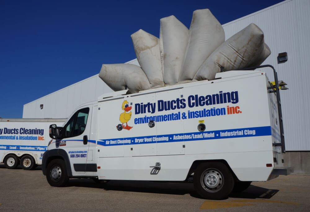 A white Dirty Ducts Cleaning van is parked outside a large building, showcasing duct cleaning company branding and contact details. Large inflated ducts rest on top of the vehicle. Another similar van is partially visible in the background.