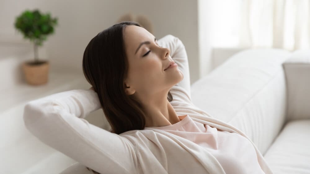 A woman with long dark hair sits back on a white couch, eyes closed and hands behind her head, exuding relaxed contentment. Soft lighting enhances the serene atmosphere, subtly guiding one's focus like effective SEO techniques, with a blurred potted plant in the background.