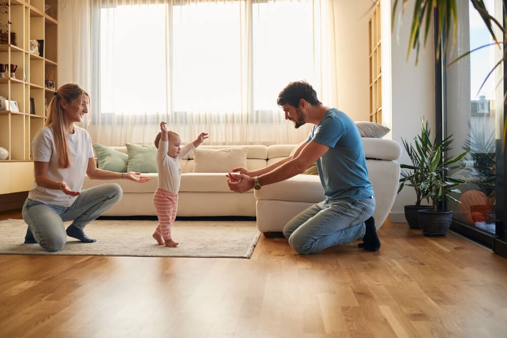 A baby takes first steps on a living room rug, supported by a smiling woman and man kneeling on either side. Sunlight streams through the windows, highlighting pristine shelves and vibrant plants—a testimony to recent mold removal ensuring a healthy environment for their little one's adventures.