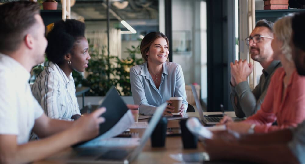 A group of people sit around a table in a modern office, engaged in a lively discussion about vermiculite removal. Laptops and papers are scattered before them. A woman in the center smiles, holding a coffee cup. The atmosphere is collaborative and friendly.