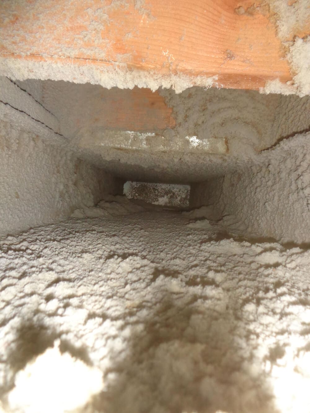 A close-up view down an air duct filled with dense dust and debris, surrounded by wooden framing, highlights the crucial need for duct cleaning. The dust accumulates along the surface, signaling compromised air quality and a call for HVAC maintenance.