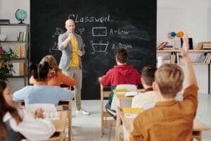 In a safe, asbestos free school environment, a teacher stands in front of a blackboard displaying math formulas, engaging with students. One raises a hand while the shelves behind show books and a globe, reflecting the commitment to education.